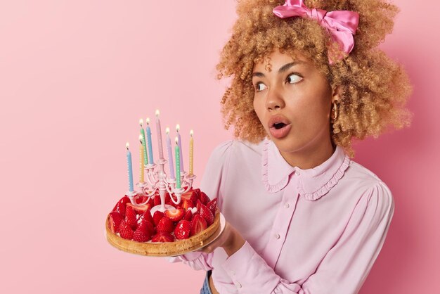 Surprised curly haired european woman holds delicious\
strawberry cake with candles makes wish on birthday wears blouse\
and bowtie looks amazed away isolated over pink background blank\
space