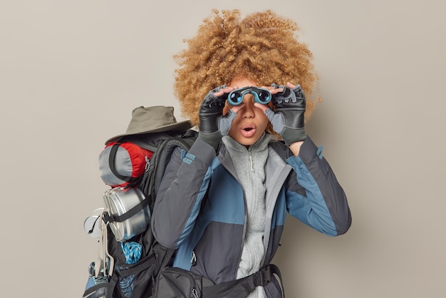 Surprised curly haired woman discovers something during hiking\
trip holds binoculars over eyes dressed in casual clothes carries\
backpack with necessary things isolated over grey background