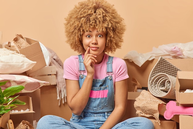 Surprised curly haired woman dressed in casual clothes\
relocates to new house or apartment poses around cardboard boxes\
with belongings thinks about mortgage goals or how to celebrate\
moving day