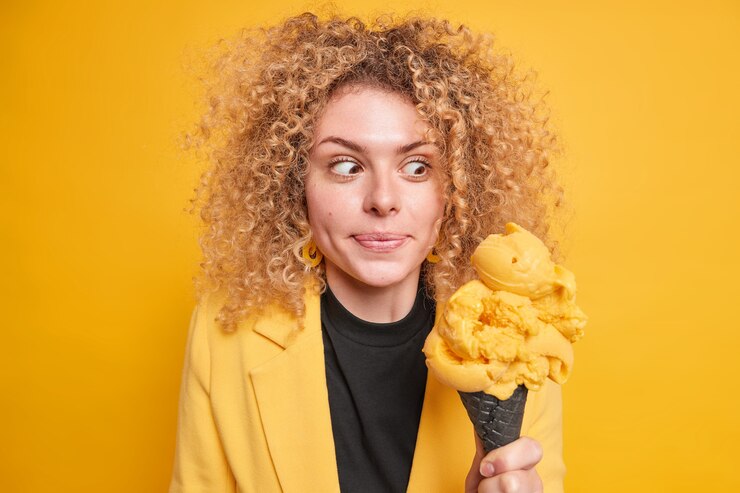 Surprised curly haired woman looks at appetizing ice cream feels temptation to eat high calorie dessert dressed in stylish clothes has walk during summer day isolated over yellow wall.