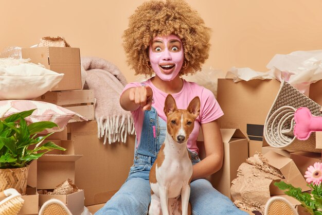 Surprised curly haired woman points directly at camera notices
something strange poses with pedigree dog dressed casually
appliesbeauty facial mask relocates to new house rents flat for
living