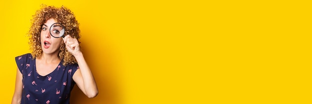 Surprised curly young woman looks at the camera through a magnifying glass on a yellow background Banner