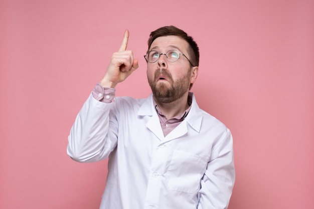 Surprised doctor in a white coat and glasses points his finger up and looks puzzled Pink background