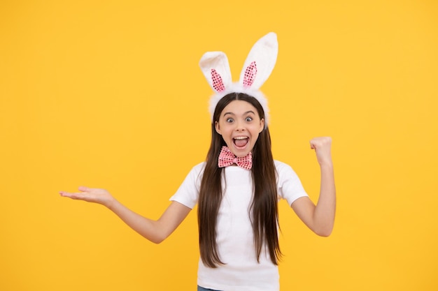 surprised easter kid girl in bunny ears and bow tie presenting product or shopping sales, copy space, easter shopping.