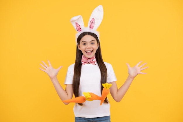 Удивленная пасхальная девочка-подросток в кроличьих ушах и галстуке-бабочке держит морковную пасху