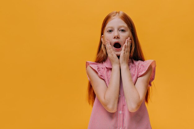 Il bambino emotivo sorpreso guarda nella fotocamera bambina rossa in camicia a righe apre la bocca e tocca le guance la ragazza posa su sfondo arancione