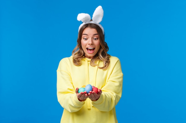 Surprised emotive cute blond caucasian girl playing holiday game, found Easter eggs look amused, wearing rabbit ears as celebrating religious holiday on sunday, 