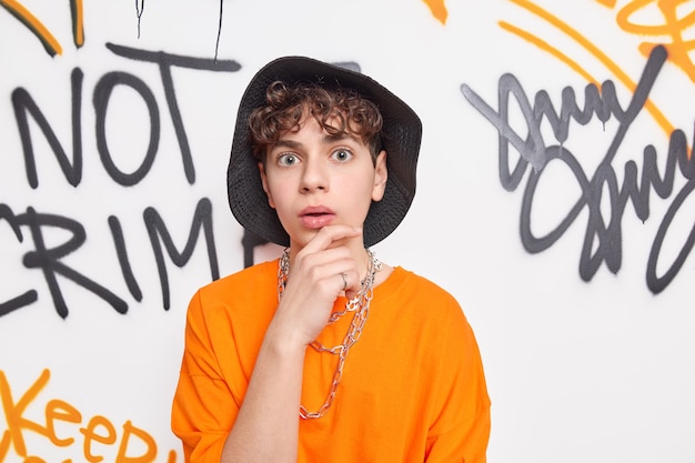 Surprised European guy holds chin stares bugged eyes at camera wears hat and orange t shirt poses against graffiti on public space wall