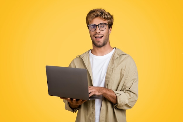 Удивленный европейский мужчина с ноутбуком смотрит на камеру на желтом фоне