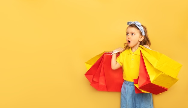 Surprised excited child holding package bags with purchases isolated on yellow wall