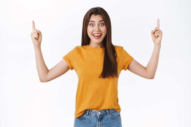 Surprised, excited and happy young smiling woman in yellow t-shirt winning amazing giveaway, showing friends and recommend try luck, pointing fingers up and gazing camera thrilled