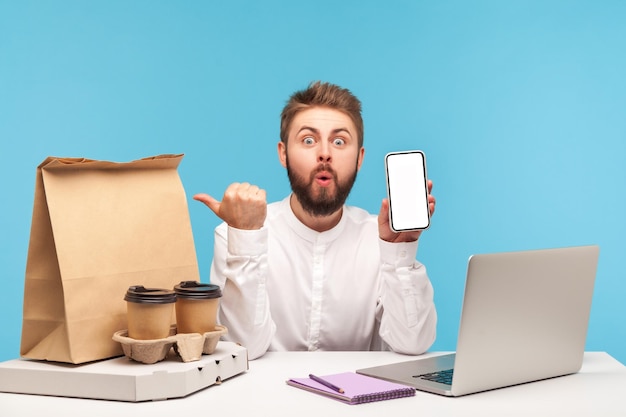 Surprised excited man pointing at fast food order on his workplace desk with thumbs and holding smartphone with empty display using app making order Indoor studio shot isolated on blue background