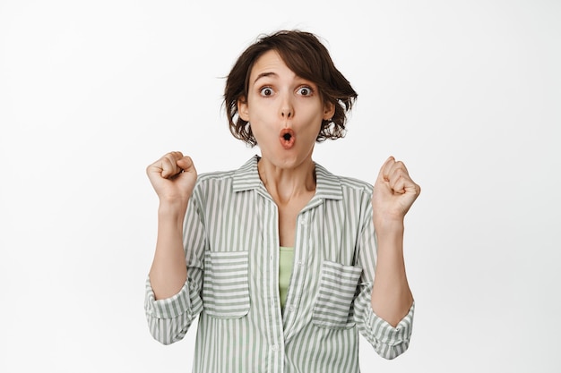 Surprised and excited woman rooting, rejoicing from win, wow face, jumping amazed, hear great news, celebrating, standing in shirt on white