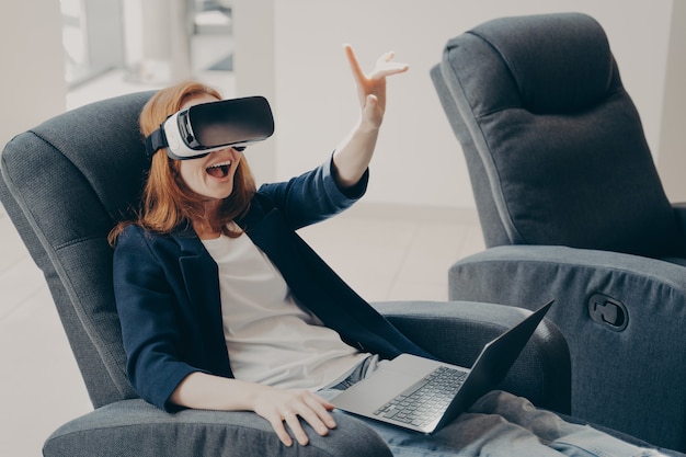 Surprised excited woman in vr headset goggles and laptop sitting in armchair in office