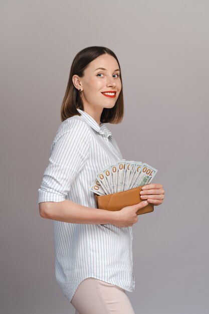 Surprised excited young woman isolated over gray wall holding money in a wallet