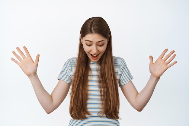 Surprised and excited young woman looking down with raised hands smiling amazed checking out something below standing against white background