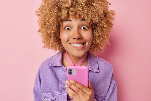 Surprised excited young woman with curly bushy hair bites lips\
looks amazed at camera holds modern cellular types text messages\
wears purple jacket isolated over pink background human\
reactions