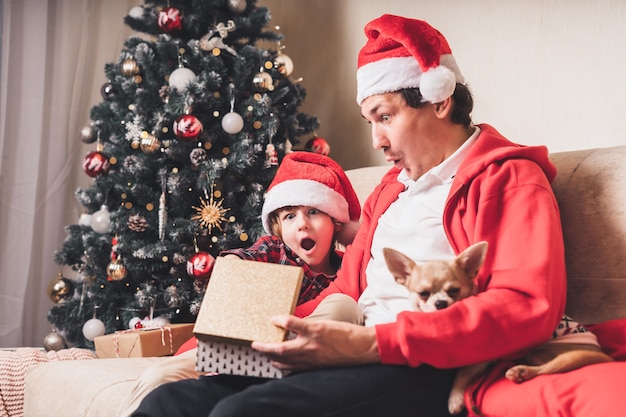 サンタの帽子で驚いた家族、父と子の男の子が自宅でクリスマスプレゼントを開きます