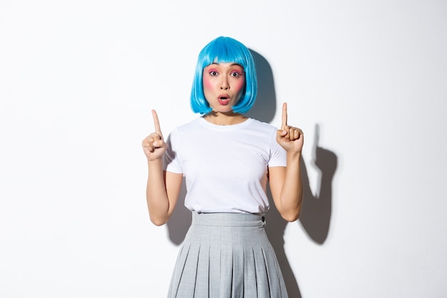 Surprised and fascinated asian girl in blue short wig, kawaii makeup, looking impressed and pointing fingers up, showing logo or promo banner, standing.