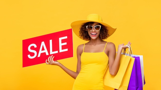 Surprised fashionable curly hair woman carrying shopping bags with red sale sign on yellow background