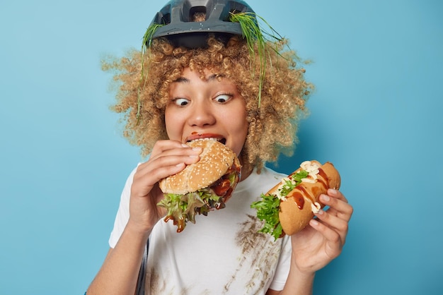 Surprised female cyclist eats delicious sandwich holds hot dog has quick snack after riding bicycle wears protective helmet with stuck grass dirty white t shirt isolated over blue background