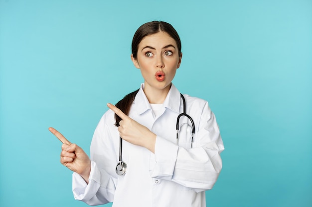 Surprised female doctor, physician with stethoscope, pointing and looking left with amazed, wow face, standing over torquoise background.