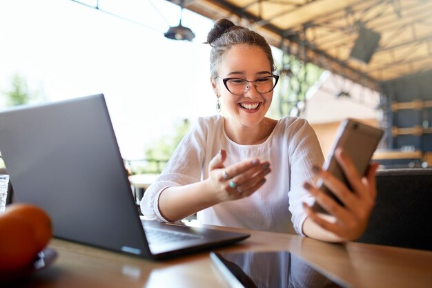 写真 スマートフォンを見ている女性は宝くじを勝ち取ったことを信じられず驚きを覚えています ビデオコールでコミュニケーションをとるアジア系白人ビジネスウーマン