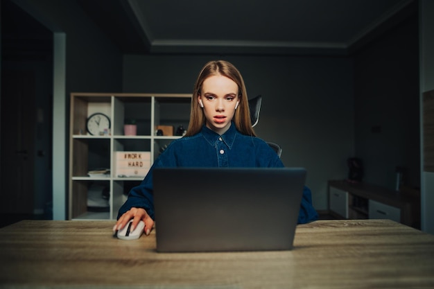 Donna freelance sorpresa in cuffia che lavora al computer portatile a casa al tavolo