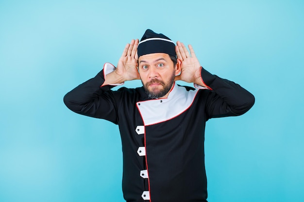 Surprised funny chef is posing to camera by holding ahnds behind ears on blue background