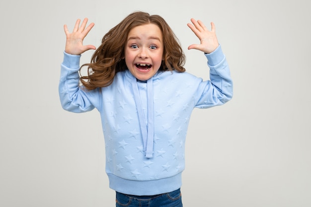 Photo surprised girl in a blue hoodie joyfully smiles while holding her hands in front of the camera on a light gray wall