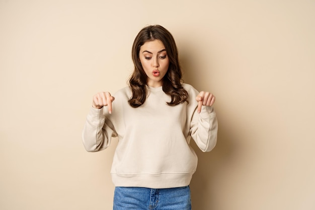 Surprised girl pointing and looking down at smth curious below beige background