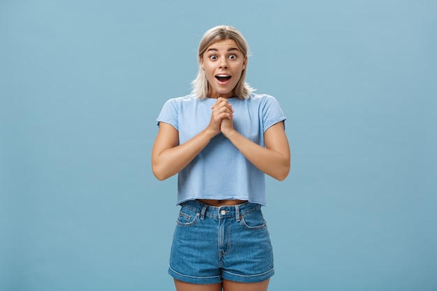 Surprised girl receiving bouquet of flowers from secret fan holding hands together on breast gasping opening mouth from amazement and happiness popping eyes from astonishment over blue wall