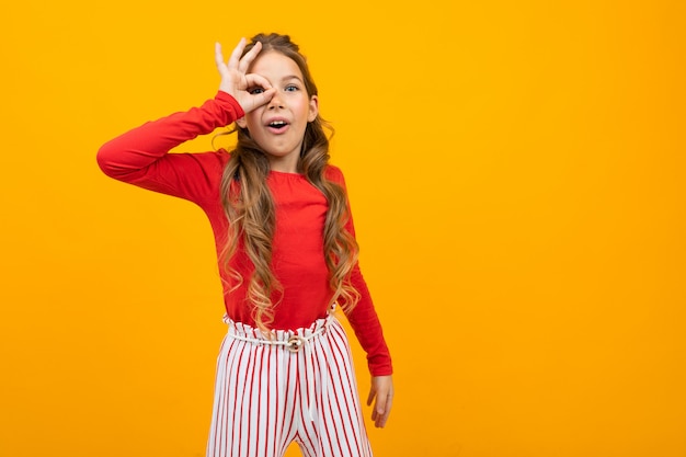 Surprised girl with curly hair in a red blouse and striped trousers peers through the palms onn orange with copy space