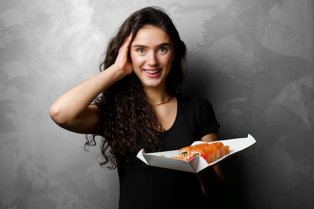 Surprised girl with sushi set philadelphia rolls in a paper box happy girl holding on a gray background. Food delivery.