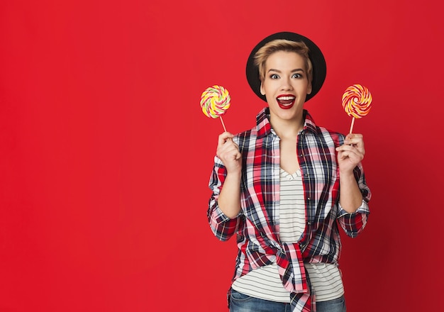 Surprised girl with two striped candies. Studio portrait at red wall, copy space