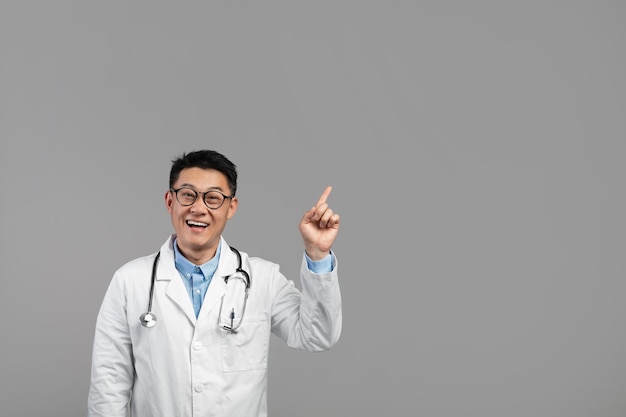 Surprised glad attractive mature asian doctor in white coat glasses with stethoscope showing finger up on empty space