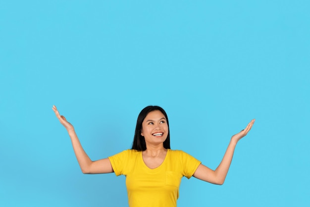 Surprised glad inspired young japanese lady in yellow tshirt raises hands up holds empty space