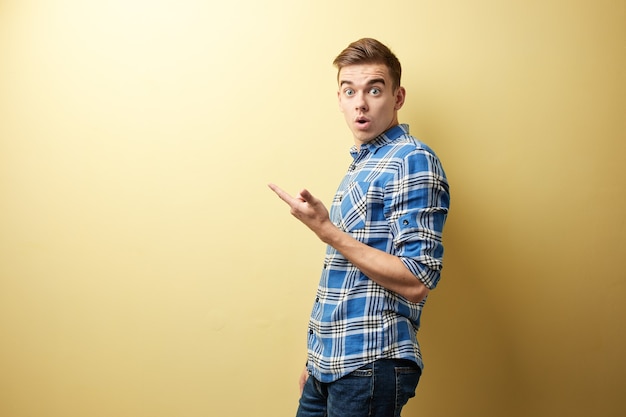 Surprised guy dressed in a plaid shirt and jeans stands next to yellow wall in the studio .