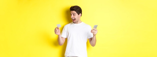 Surprised guy holding smartphone and credit card online shopping on black friday standing over yello