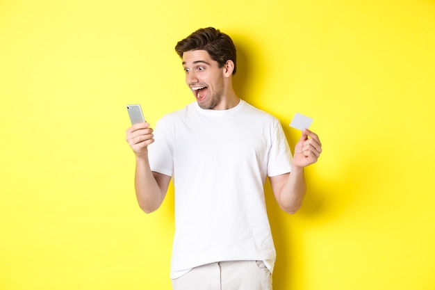 Surprised guy holding smartphone and credit card, online shopping on black friday, standing over yellow background.