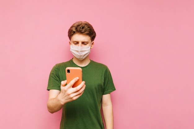 의료용 마스크와 녹색 티셔츠를 입은 놀란 남자는 스마트폰에서 인터넷을 사용하고 코로나바이러스 코로나바이러스 전염병 격리 covid19 핑크 배경에 대한 뉴스를 읽습니다.