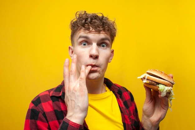 surprised guy student eats big tasty burger on yellow background and licks the sauce on his finger