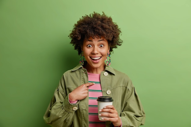 Sorpresa felice donna afroamericana indica se stessa chiede se scegli me non può credere in qualcosa che indossa abiti alla moda beve caffè da asporto isolato sopra il muro verde