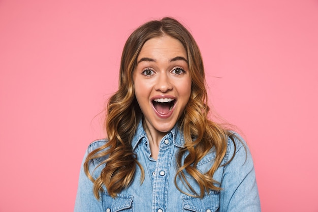 Surprised happy blonde woman wearing in denim shirt rejoices and looking at the front over pink wall