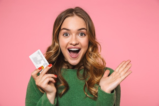 Donna bionda felice sorpresa che indossa in maglione verde che tiene la carta di credito e che guarda la parte anteriore sopra il muro rosa