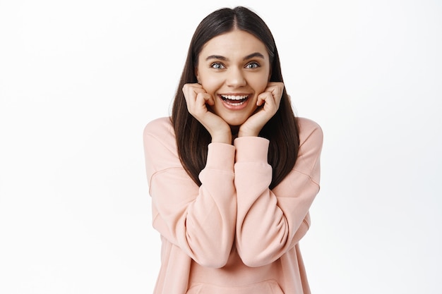 Surprised and happy brunette girl gasping amazed, smiling and holding hands on cheeks, looking at something awesome, receiving a gift or pleasant news, white wall