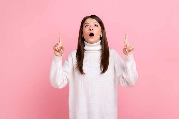 Surprised happy brunette girl looking astonished and pointing up copy space, showing empty place, wearing white casual style sweater. Indoor studio shot isolated on pink background.