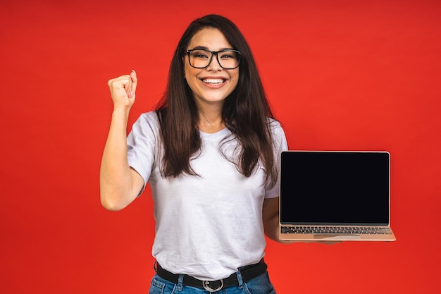 Surprised happy brunette woman in casual showing blank laptop computer screen while looking at the camera isolated over red background