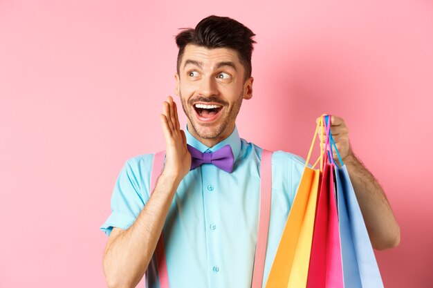 Surprised happy man screaming of joy, looking aside at logo and holding shopping bags, standing over pink background.