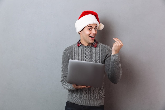 Удивленный счастливый человек в свитере и рождественской шляпе держит портативный компьютер и смотрит